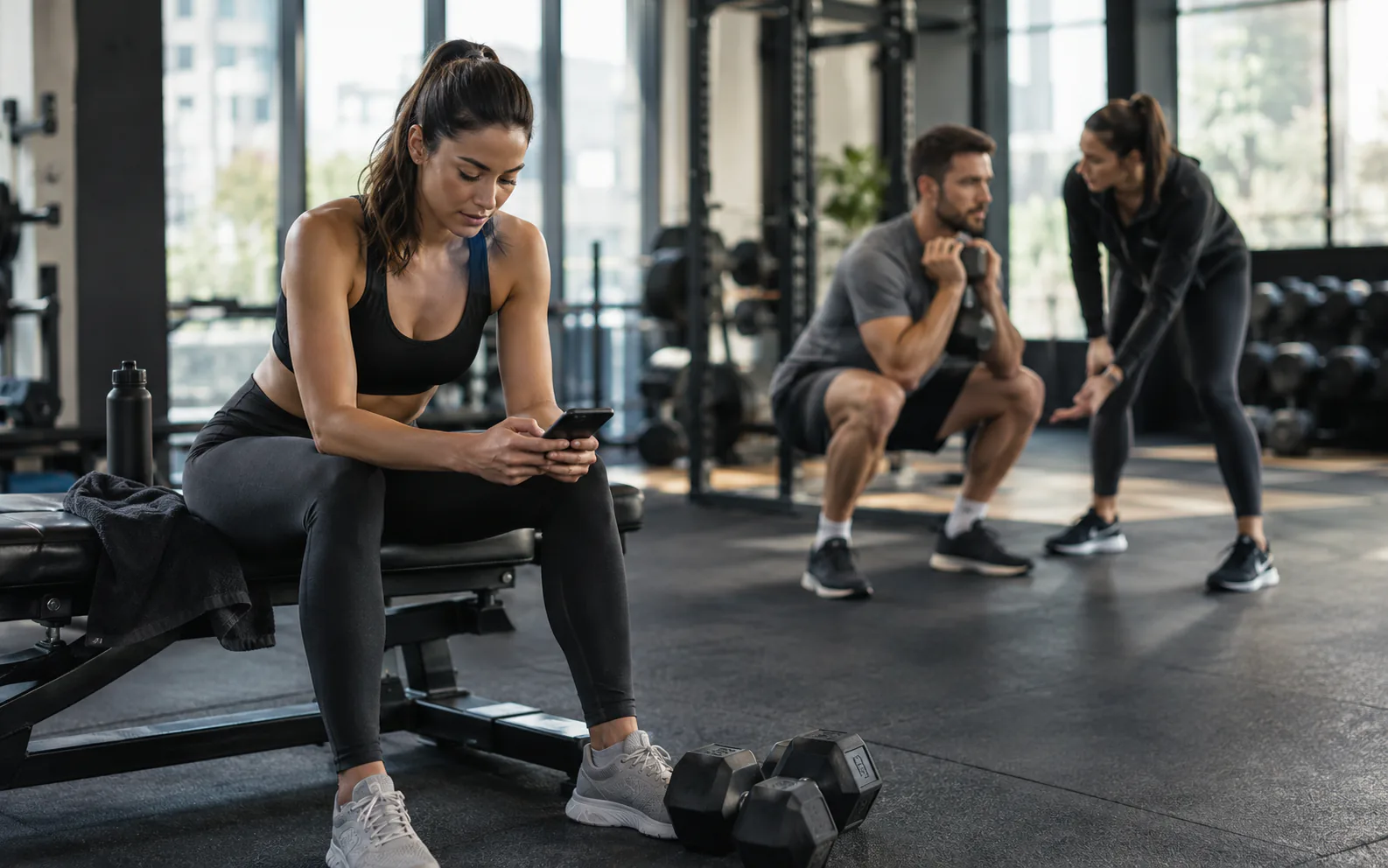 Frau trägt ihr Training im Handy ein, während hinter ihr ein Personal Trainer einen Kunden durch einen Goblet Squat begleitet